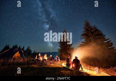 Night camping in mountains under starry sky. Group of people hikers having a rest near campsite, burning campfire and tourist tent. Concept of tourism, hiking and adventure. Stock Photo