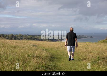 A man walks along the top of a hill on a grass path. He looks out over the sea and land in the distance. It looks relaxing and inviting for a hike. Stock Photo