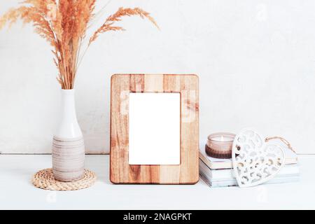 Mockup wooden picture frame, books, aroma candle, carved decor heart and pampas grass in vase on white table. Copy space. Stock Photo