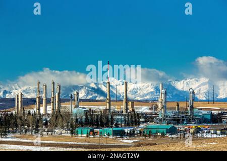 Gas plant in the winter with snow-covered Rocky mountains in the background with blue sky and clouds over the mountains, North of Cochrane Stock Photo