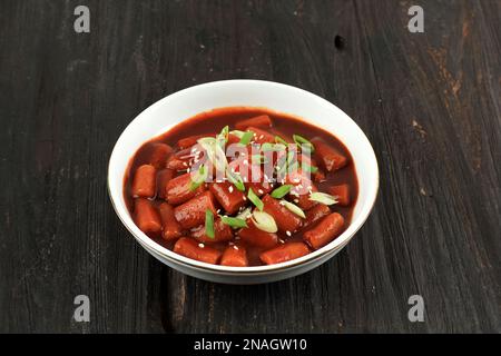 Tteokbokki, Korean RIce Cake with Red Spicy Gochujang Sauce, Popular Street Food from South Korea. Served on White Plate above Black Wooden Table Stock Photo