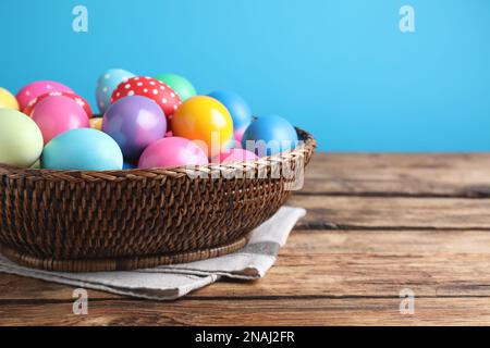 Colorful Easter eggs in wicker basket on wooden table against light blue background, closeup. Space for text Stock Photo