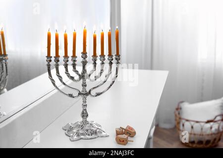 Silver menorah near dreidels with symbols He, Pe, Nun, Gimel on white console table in room decorated for Christmas Stock Photo