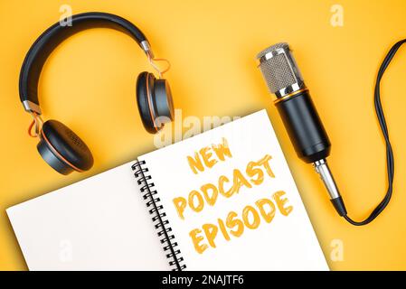 NEW PODCAST EPISODE text on notepad next to headphones and recording microphone, podcasting concept on orange background Stock Photo