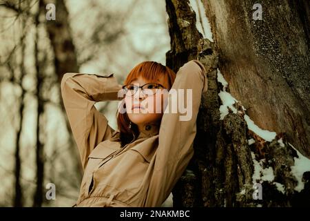 Portrait of a beautiful ginger Asian woman in a beige dress leaning against a thick snow-covered tree trunk in a winter park. Outdoors. Stock Photo