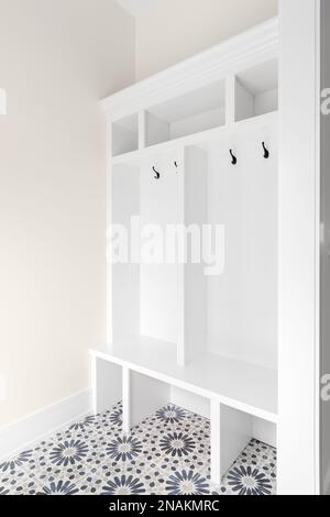 A white bench in a mudroom foyer with a bench seat, coat hooks, shelving, and a patterned tile floor. Stock Photo