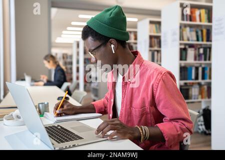 Focused African guy online student taking notes during webinar, studying in library on laptop Stock Photo