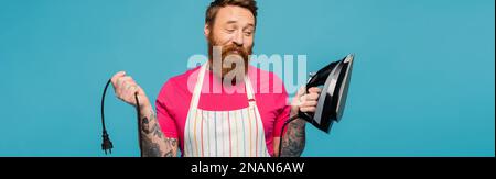 smiling and skeptical man in striped apron holding electric iron isolated on blue, banner,stock image Stock Photo