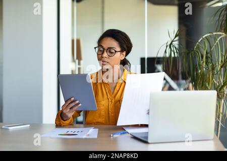 Serious concentrated black businesswoman analyzing data in documents and tablet, sitting at workplace in office interior Stock Photo