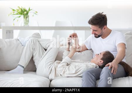 Nice young couple sitting on the white couch in the cozy room while watching something on the cell phone device and talking at home. Indoors Stock Photo