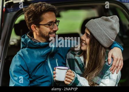Satisfied millennial caucasian couple sitting in trunk of car in cold forest, enjoy adventure and cup of hot drink Stock Photo
