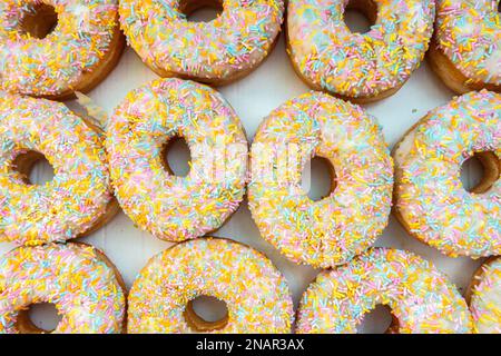 A box of glazed ring doughnuts with white frosting and colourful sprinkles Stock Photo