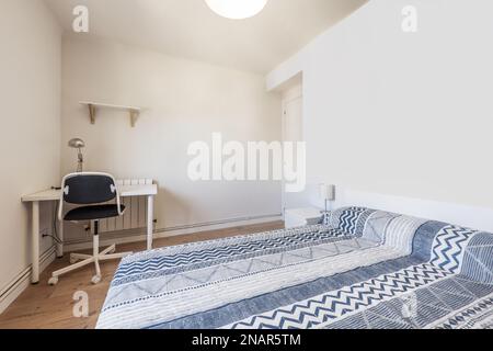 A bedroom with a double bed with a blue patterned bedspread, a white desk with a swivel chair with chestnut hardwood floors and a white wooden front d Stock Photo
