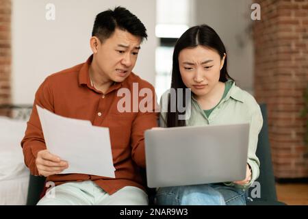 Angry asian spouses checking bills and using laptop, unhappy middle aged man and young woman counting monthly spendings Stock Photo