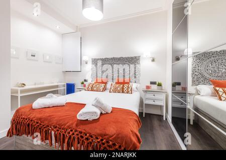bedroom with a double bed with orange cushions and a gray wooden headboard, different bedside tables and a wardrobe with white sliding doors with a mi Stock Photo