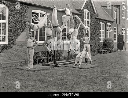 A gymnastic display outdoors with 10 men posing on a set of parallel bars, UK c.1900. Balance skills plus strength, fitness and flexibility are called upon. The man in the top position is supported with his feet is on the necks of the two gymnasts beneath him who are in the handstand position. The gymnast at the front of the group is in the ‘bridge’ position. The long exposure means there is some motion blur caused by movement by some of the men holding the pose – a vintage 1800s/1900s, Victorian/Edwardian photograph. Stock Photo