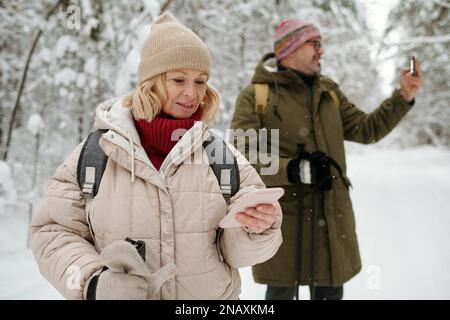 Two mature backpackers using their smartphones during winter trip on weekend while standing in pinetree forest covered with snow Stock Photo