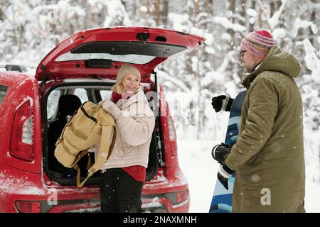 Happy senior woman with backpack looking at her husband with snowboard while taking baggage out of car trunk Stock Photo