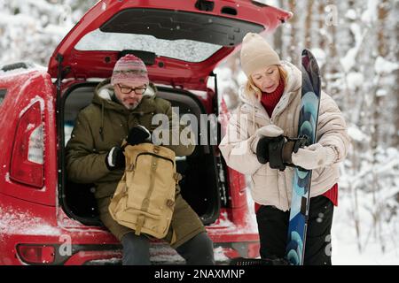 Mature blond woman looking at snowboard during winter trip while her husband sitting in hatchback car trunk and unzipping backpack Stock Photo