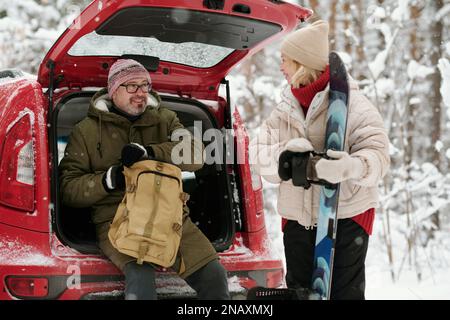 Mature active woman with snowboard looking at her husband unzipping backpack while sitting in hatchback car trunk during winter hike Stock Photo