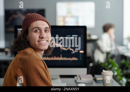 Happy young IT support manager looking at camera while sitting by workplace in front of computer screen with graphic data Stock Photo