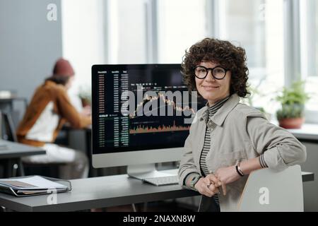 Young smiling businesswoman or crypto analyst in casualwear sitting by workplace with computer monitor and graphic data on screen Stock Photo