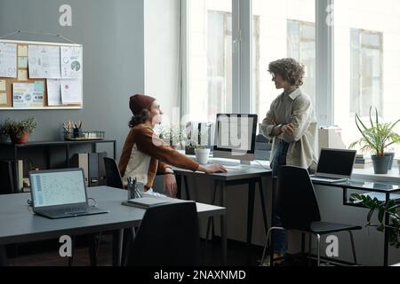 Two young intercultural analysts communicating at meeting in office while one of them sitting by desk in front of computer monitor Stock Photo