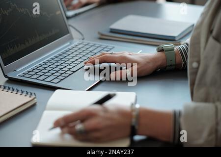 Close-up of young female economist keeping hand over laptop keyboard and making notes in notepad while organizing work or analyzing data Stock Photo