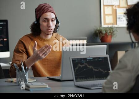 Young confident male operator of helpdesk or IT support manager with headset answering questions of online clients in front of laptop Stock Photo