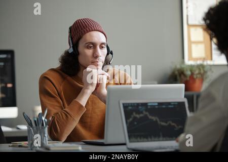 Young serious male IT support operator sitting by workplace in front of laptop and looking at camera while listening to online client Stock Photo