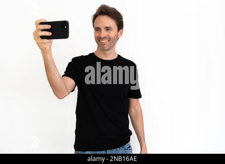 Handsome male model is posing in a black t-shirt taking a selfie and looking happy. Black mock-up. Stock Photo