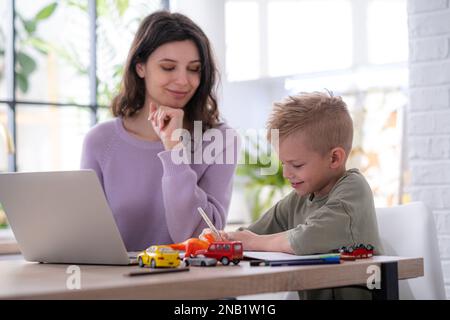 Work from home and family education, Mom and son using remote access technology for learning and work, woman works on laptop, child writes in textbook Stock Photo
