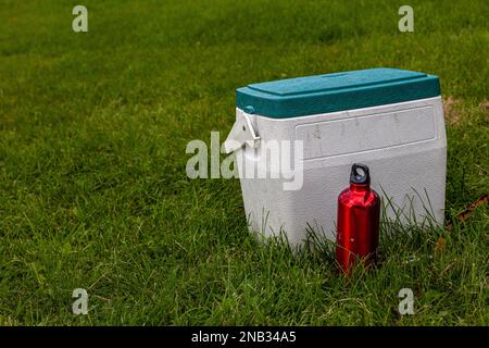 White cooler box with green lid and a red water bottle on a green lawn Stock Photo