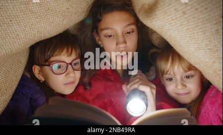 Teen girl and toddler child sisters under blanket plaid covers with flashlight reading interesting fairytale story book using flashlight. Female three siblings children kids friends in wigwam at home Stock Photo