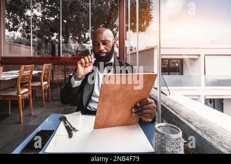 A bald black man dressed in a sleek black tuxedo and bow tie is perusing the cork color menu at a restaurant, located on a rooftop, with a side view w Stock Photo