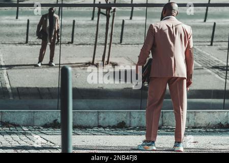 A shot of a balding back faced stylish black man in a cream-colored suit with a briefcase in his hand and trendy sneakers, waiting at the crosswalk ha Stock Photo