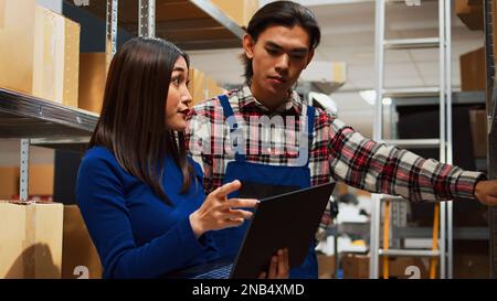 Warehouse employees working on inventory with laptop, walking around storage room racks with carton boxes. Entrepreneur and worker planning stock distribution, small business concept. Stock Photo