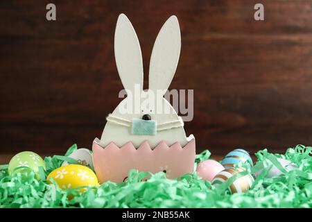 Cute bunny figure in protective mask and dyed eggs on wooden background. Easter holiday during COVID-19 quarantine Stock Photo