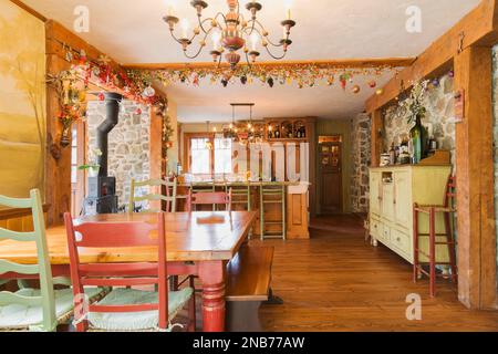 Dining room with antique pine wood table, red stained wooden legs, wood and woven rawhide seat chairs plus kitchen in background in old 1826 home. Stock Photo