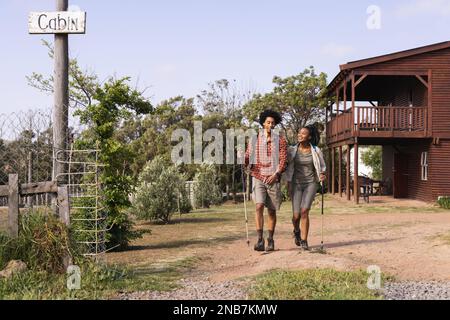 Happy african american couple walking with hiking sticks in countryside on sunny day. Country, nature and lifestyle concept. Stock Photo
