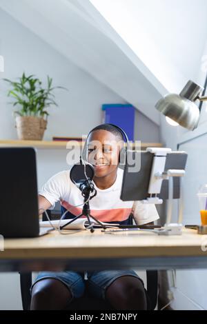 Vertical image of happy african american teenager with headphones and microphone recording podcast. Teenager, adolescence, hobby and spending free tim Stock Photo