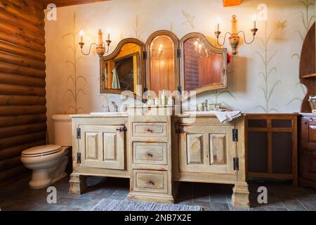 Bleached and distressed pinewood cabinets, antique wall-mounted folding mirror and white porcelain toilet in main bathroom inside old 1826 home. Stock Photo