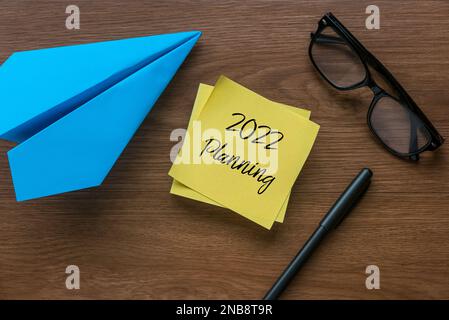 2022 planning written on yellow memo note with paper plane, pen and glasses on wooden background. Stock Photo