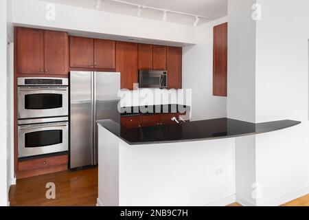 A small kitchen with cherry wood cabinets, stainless steel appliances, and black granite countertops. Stock Photo