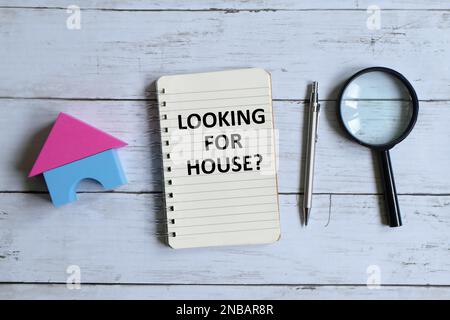 Top view of house model,pen,magnifying glass and notebook written with 'LOOKING FOR HOUSE?' on white wooden background. Stock Photo
