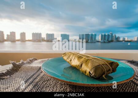 Wrapped Hallaca or Tamale over a rustic table and a blue dish, Mexican and Venezuelan traditional food with a Miami, Florida background Stock Photo