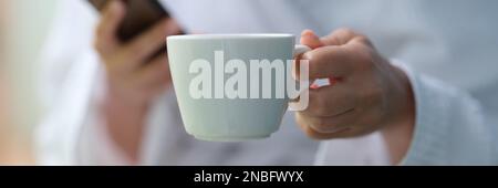 Cropped photo of a woman in a terry bathrobe holding mobile phone and cup Stock Photo