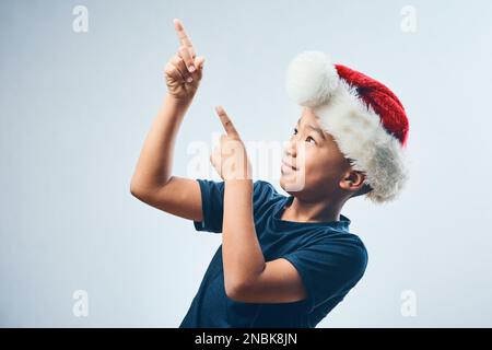 I think I spot Santas sleigh. Studio shot of a cute little boy wearing a Santa Claus hat and pointing against a grey background. Stock Photo