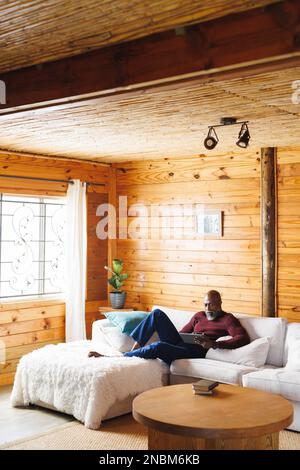 Bald african american senior man using digital tablet while relaxing on couch at log cabin. Copy space, unaltered, retirement, vacation, solitude, hol Stock Photo