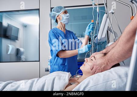 Two doctors checking on unconscious woman patient at intensive care unit and monitoring her health with medical equipment and sensors. ICU Stock Photo